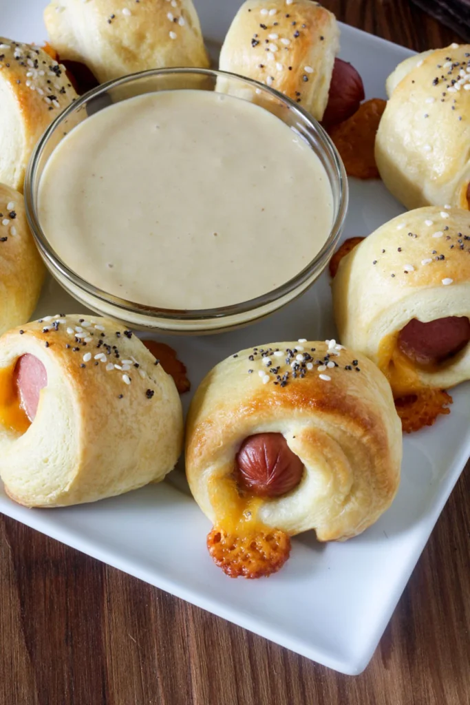 Pigs in a blanket on white plate with dipping sauce in clear bowl in the center