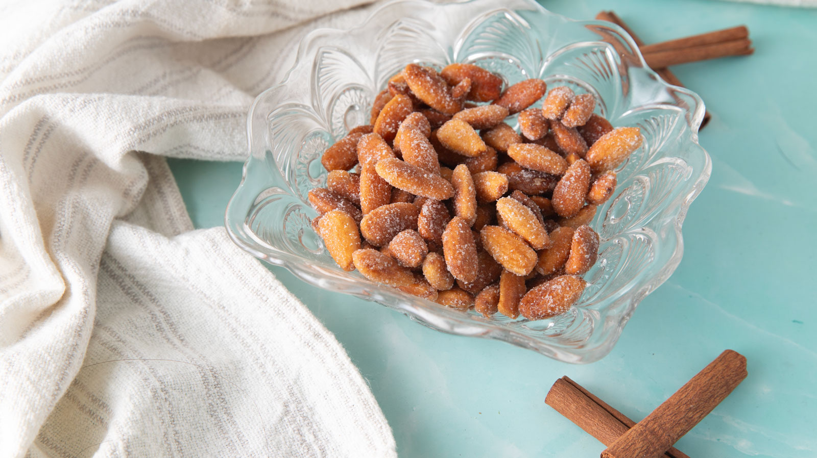 Glass bowl filled with honey roasted almonds with cinnamon sticks on a green board.