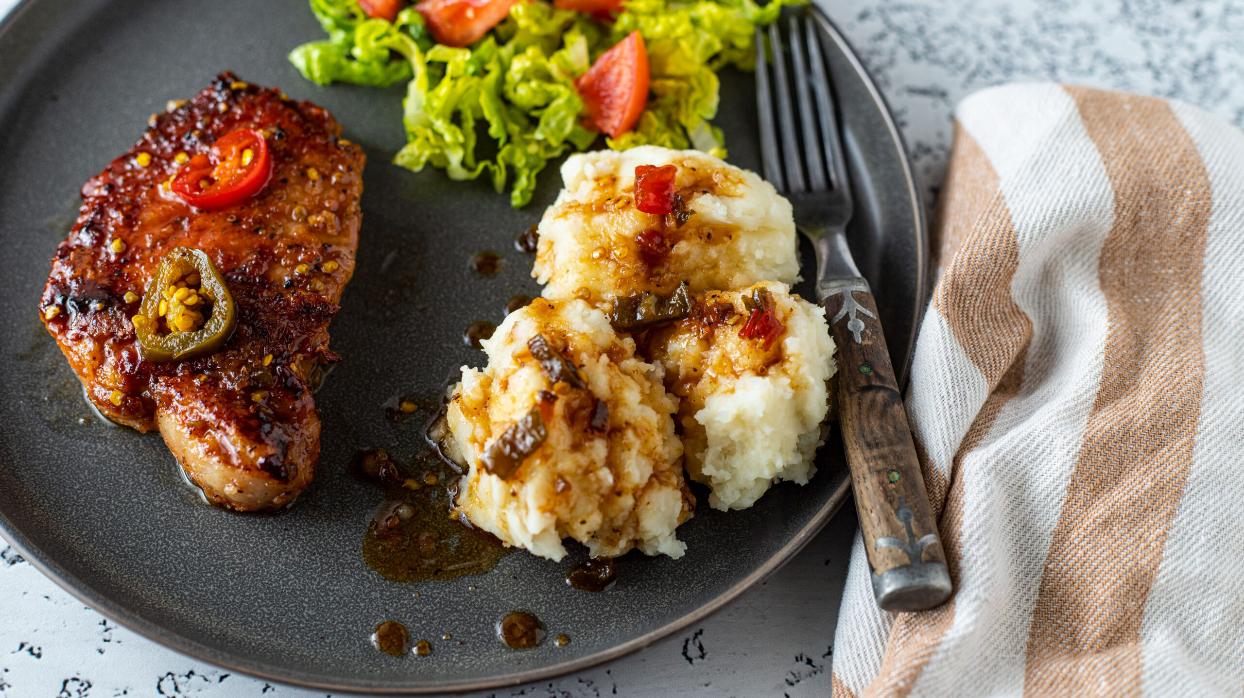 Pork chop with candied jalapeños on a gray plate with potates and salad..